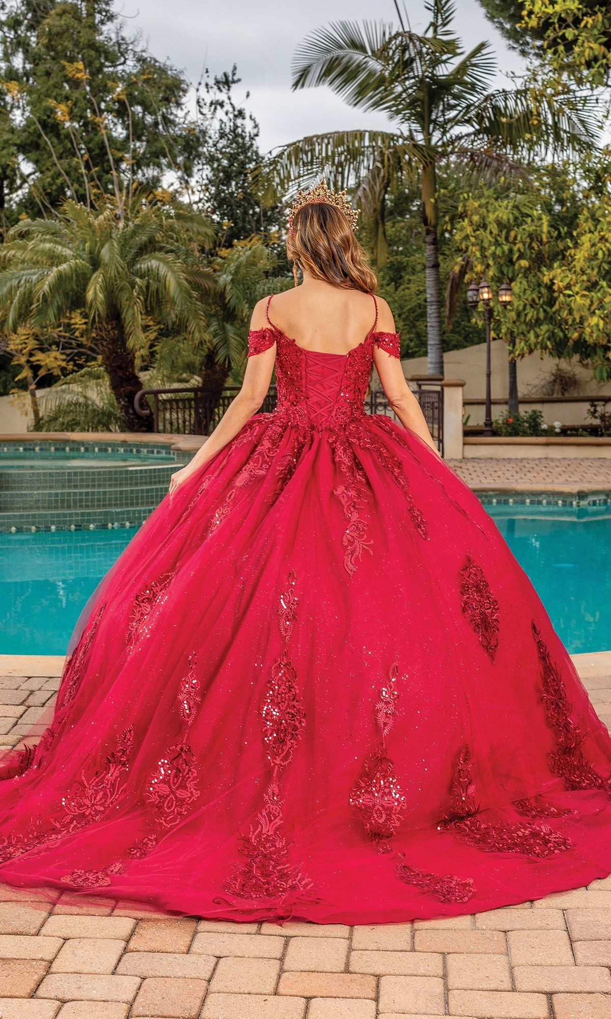A woman wearing Quinceanera Dress 1811 By Dancing Queen, an off-the-shoulder embellished burgundy gown, stands with her back to the camera by a pool amid palm trees and lush greenery.